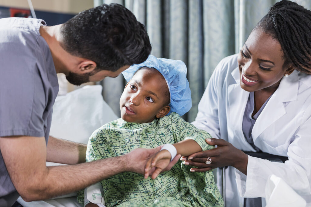 Una niña afroamericana de 6 años de edad sentada en la camilla de un hospital, con una doctora y un enfermero que la atienden.   Lleva bata hospitalaria y gorro quirúrgico. Una enfermera en ropa quirúrgica, con un joven de Medio Oriente de unos veintitantos años, de pie a un lado y una doctora, una mujer afroamericana madura de unos 40 años al otro.